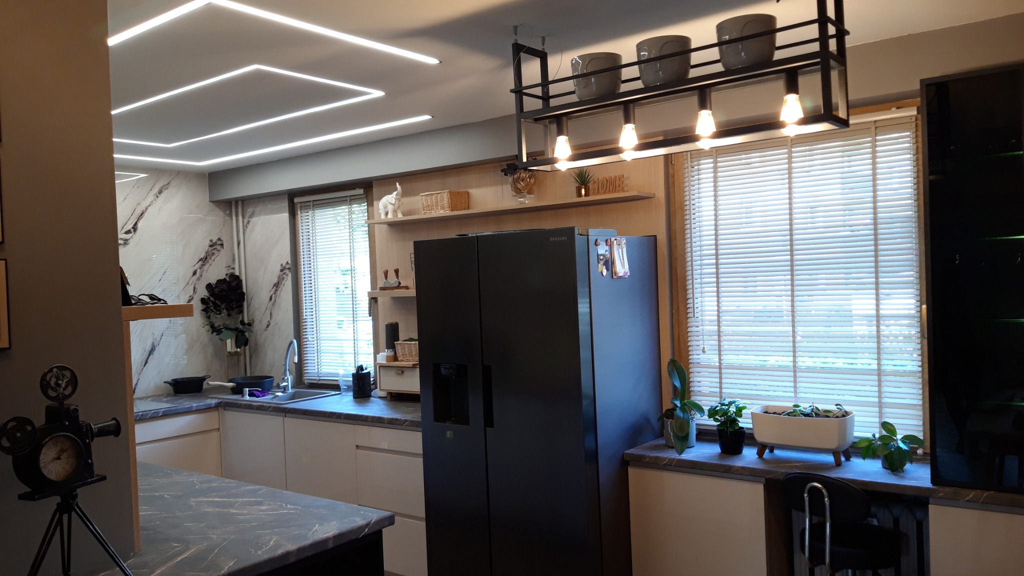 Nowoczesna kuchnia z białymi szafkami, czarną lodówką side-by-side i marmurowym blatem, oświetlona geometrycznymi lampami sufitowymi LED i wiszącą lampą z doniczkami.