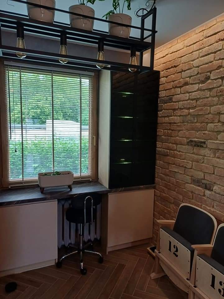 Kącik z biurkiem pod oknem, zabudowa z czarną witryną, ceglana ściana i drewniane krzesła kinowe z numerami 12 i 13.