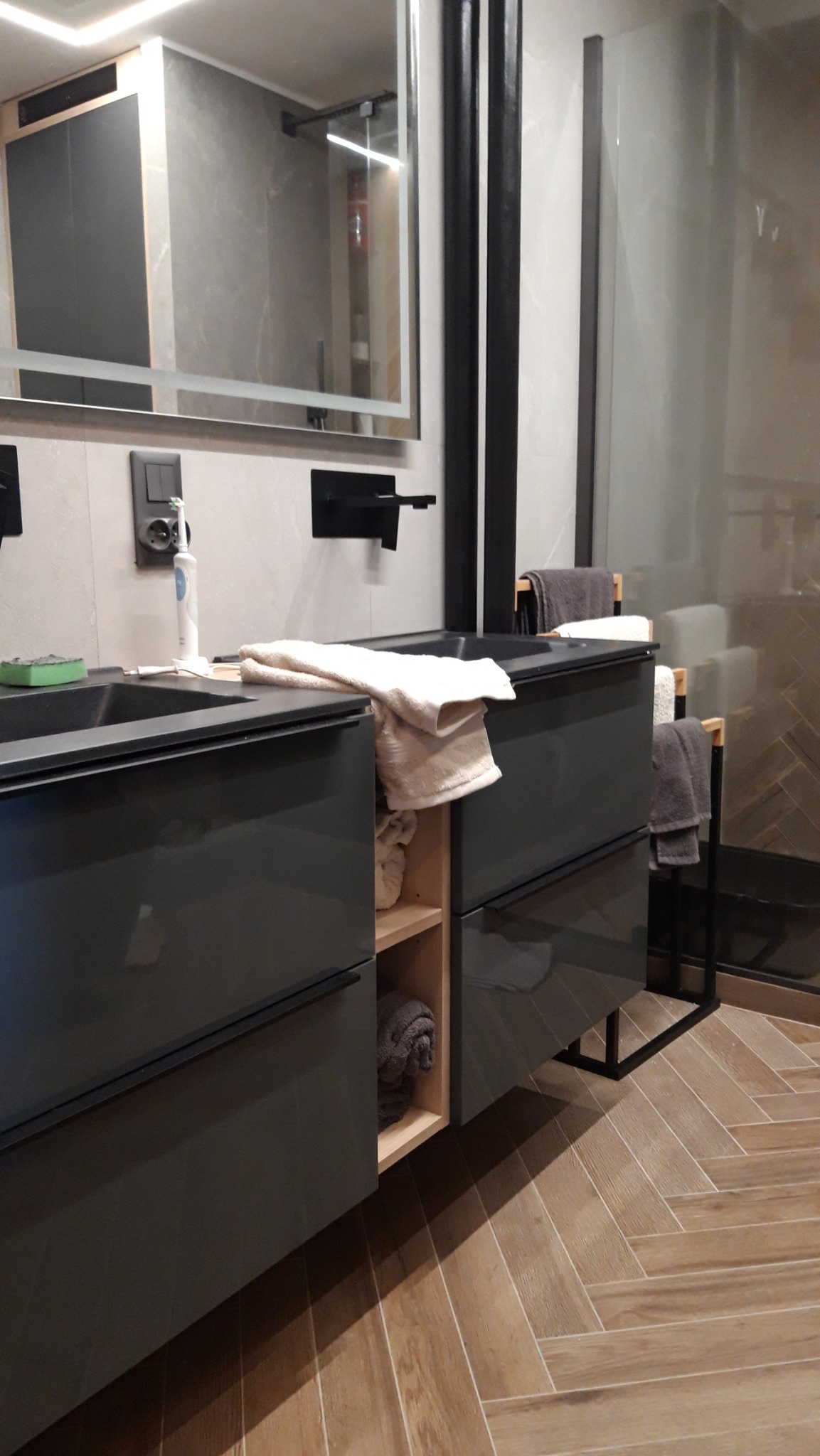 Nowoczesna szafka łazienkowa na wymiar z szufladami i otwartymi półkami na ręczniki, zrealizowana w Piotrkowie Trybunalskim, z drewnianą podłogą ułożoną w jodełkę.