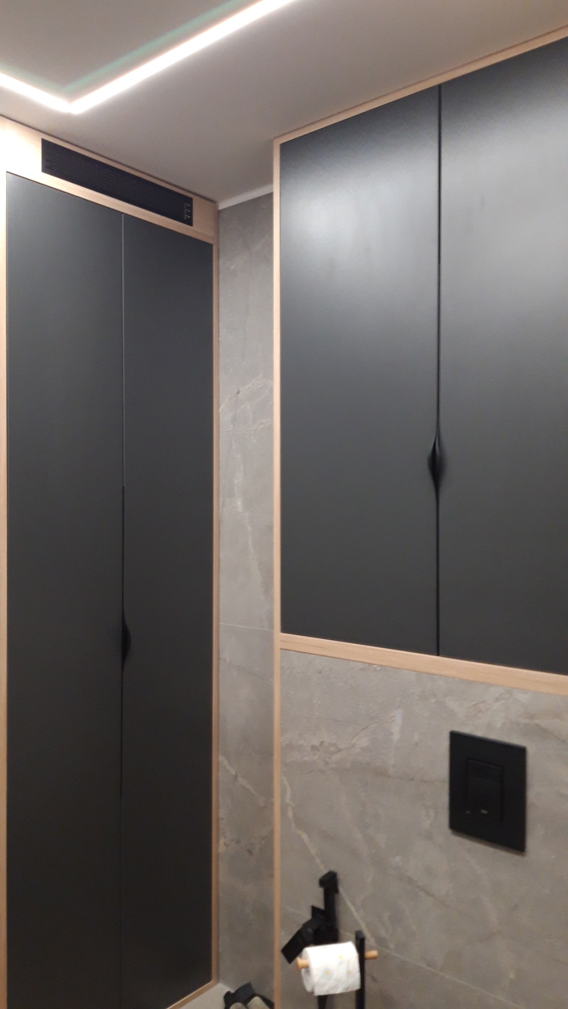 Ciemnoszare szafki z drewnianymi ramami w łazience wykończonej marmurowymi płytkami, widoczny czarny przycisk spłuczki i uchwyt na papier toaletowy.