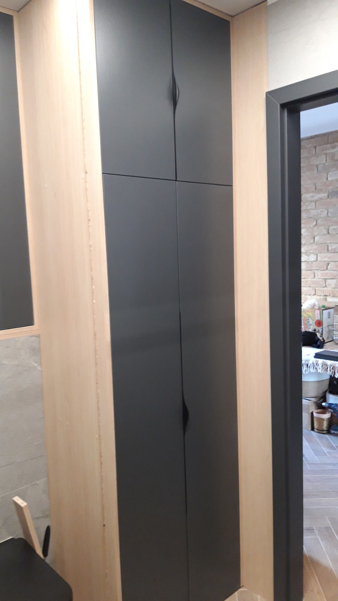 Wysoka, grafitowa szafa w zabudowie z drewnianymi bokami, z minimalistycznymi, podłużnymi uchwytami, widoczna z boku, obok wejście do pokoju z ceglaną ścianą.