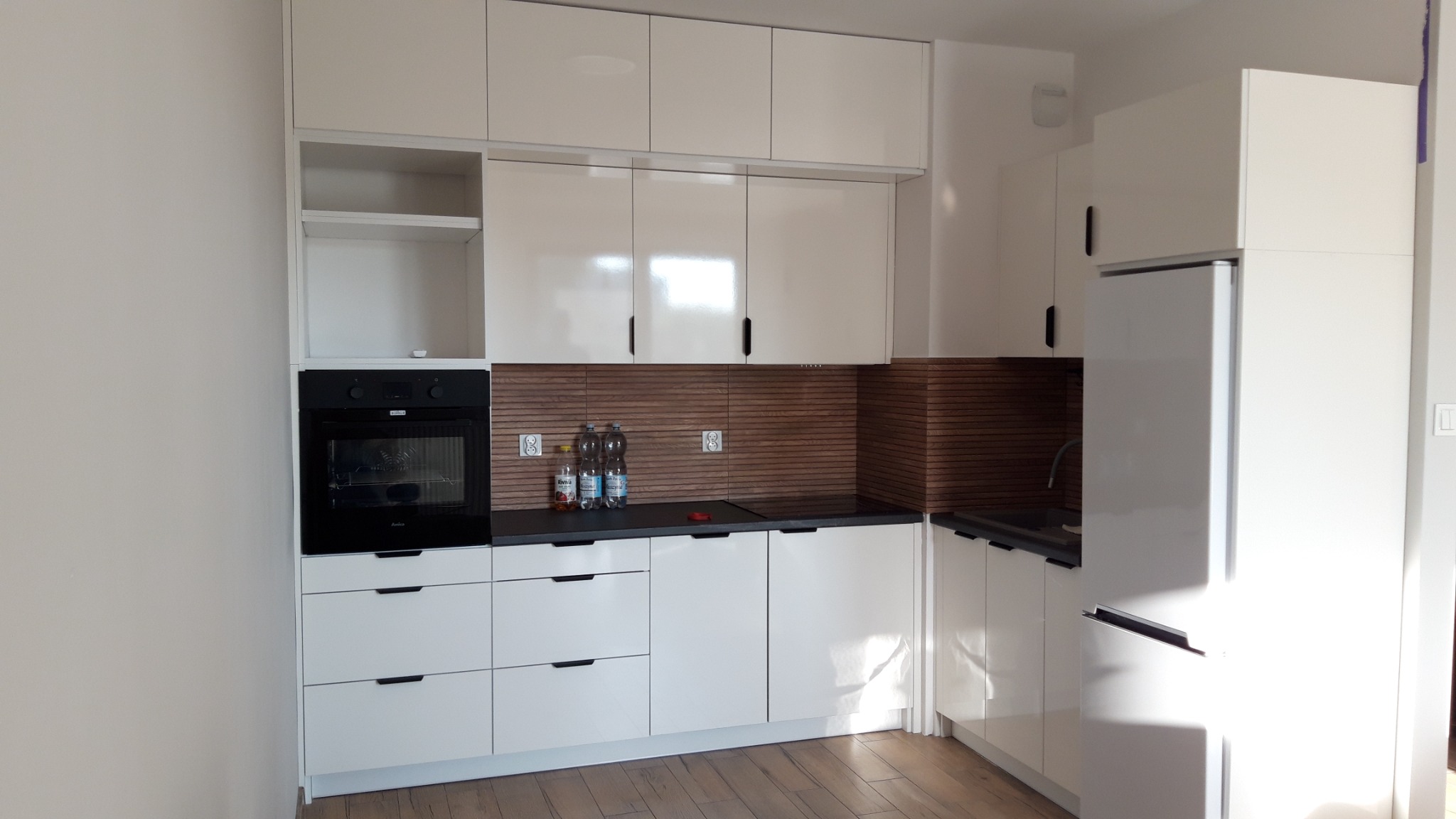 Nowoczesna, biała zabudowa kuchenna z piekarnikiem w czarnej obudowie, drewnianym panelem ściennym i czarnym blatem, uzupełniona o białą lodówkę.