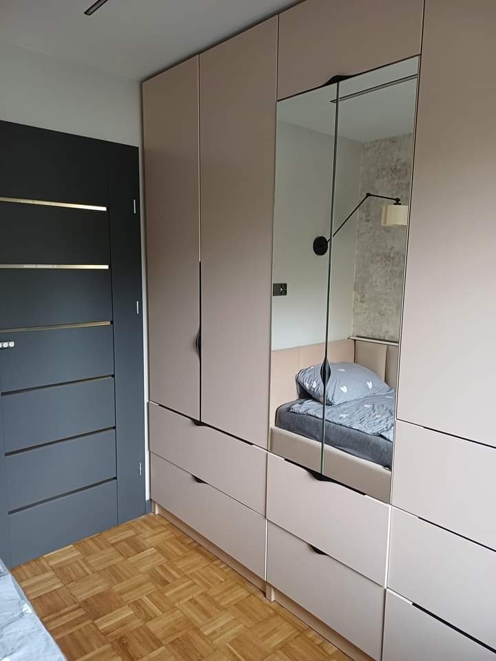 Jasna zabudowa meblowa z szafami, szufladami i lustrzanym frontem, odbijająca fragment sypialni z łóżkiem i lampką ścienną, na tle parkietu ułożonego w kwadraty oraz ciemnych drzwi z poziomymi...