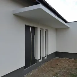 Wykończona elewacja domu jednorodzinnego z szarą stolarką okienną i drzwiową, oraz widocznym zadaszeniem nad wejściem, pomalowana na biało, z ciemnoszarą podmurówką.