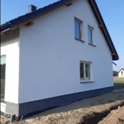 Nowoczesny dom jednorodzinny z białą elewacją, czarną podmurówką i oknami z pomarańczowymi ramkami w trakcie prac ziemnych wokół fundamentów.