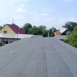 Nowo pokryty, płaski dach z papy termozgrzewalnej, widoczna butla gazowa i butelka wody, w tle zabudowania i zieleń.