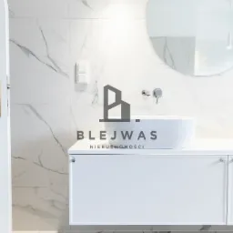 Nowoczesna łazienka z dwoma umywalkami nablatowymi, okrągłym lustrem i białymi płytkami imitującymi marmur, częściowo widoczne białe drzwi z mosiężną klamką, logo agencji nieruchomości na blacie.