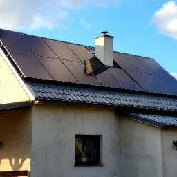 Dom jednorodzinny z zainstalowanymi panelami słonecznymi na dachu pokrytym blachodachówką, komin w kolorze elewacji, błękitne niebo z chmurami w tle.