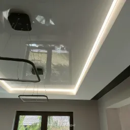 Biały, błyszczący sufit z oświetleniem LED wzdłuż krawędzi i nowoczesną lampą wiszącą z kwadratowymi elementami, odbijający widok okna z ogrodem.