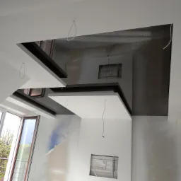 Wnętrze w trakcie prac wykończeniowych: widoczne płyty kartonowo-gipsowe na ścianach i elementach sufitu podwieszanego z wycięciami na oświetlenie, kable zwisające z sufitu, okno balkonowe w tle.