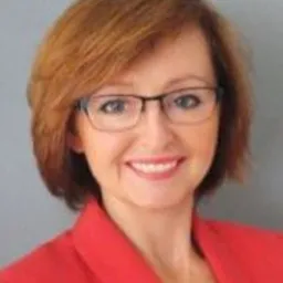 Portret kobiety w okularach, ubrana w czerwoną marynarkę, uśmiechająca się do kamery, z krótkimi, rudymi włosami.