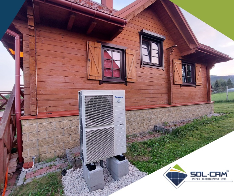 Zewnętrzna jednostka pompy ciepła Panasonic Aquarea zainstalowana przy drewnianym domu z okiennicami, na betonowych podstawach i otoczona białym żwirem.