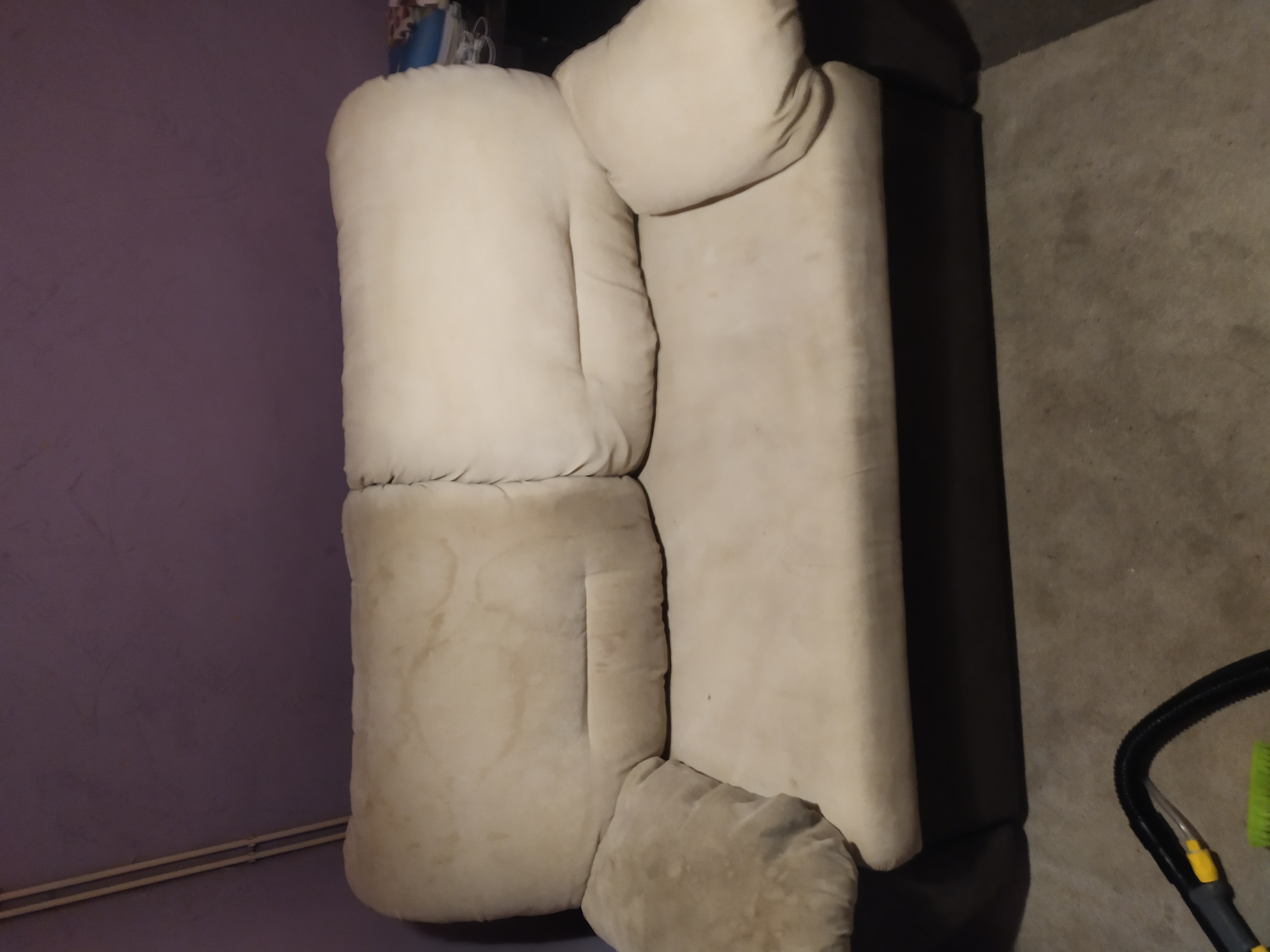 Beżowa, modułowa sofa z widocznymi plamami na siedzisku, ustawiona pod fioletową ścianą. W tle widoczny fragment odkurzacza piorącego.