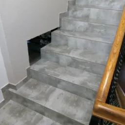 Wykończone schody wewnętrzne wyłożone szarymi płytkami imitującymi kamień, z drewnianą balustradą i dekoracyjnym oświetleniem pod schodami.