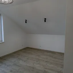 Poddasze po remoncie: jasne ściany, panele podłogowe w odcieniach szarości, okno z widokiem na drzewa, dwa czarne reflektory punktowe na skosie sufitu.