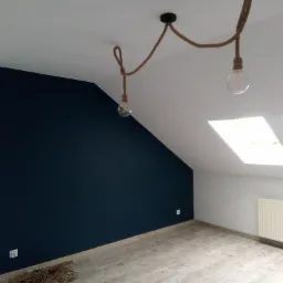 Pokój na poddaszu po remoncie, z granatową ścianą akcentującą, drewnianymi panelami podłogowymi i lampami wiszącymi na sznurze, widoczne okna dachowe i grzejnik.
