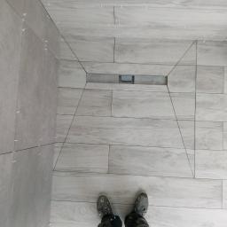 Krzysztof - Świeżo ułożone, jasnoszare płytki imitujące drewno, z widocznym odpływem liniowym i krzyżykami dystansowymi w spoinach, częściowo na ścianie, z perspektywy osoby stojącej na podłodze w ubrudzonych...