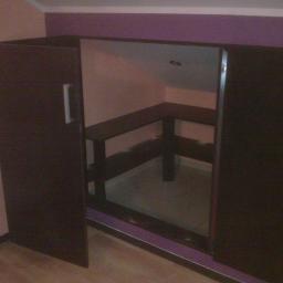 Krzysztof - Ciemnobrązowa szafa z przesuwnymi drzwiami, widoczna półka wewnątrz, na tle ściany w odcieniach różu i fioletu. Ujęcie z niskiego kąta.