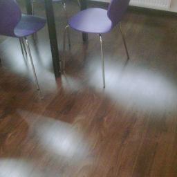 Krzysztof - Ułożone panele podłogowe w odcieniach brązu, widoczne fragmenty krzeseł z fioletowymi siedziskami i metalowymi nogami, oraz stopa w domowym obuwiu w lewym dolnym rogu kadru.