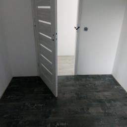 Krzysztof - Wnętrze pokoju z ciemną podłogą imitującą drewno i otwartymi, białymi drzwiami z poziomymi przeszkleniami, widok na sąsiednie pomieszczenie z jasną podłogą.