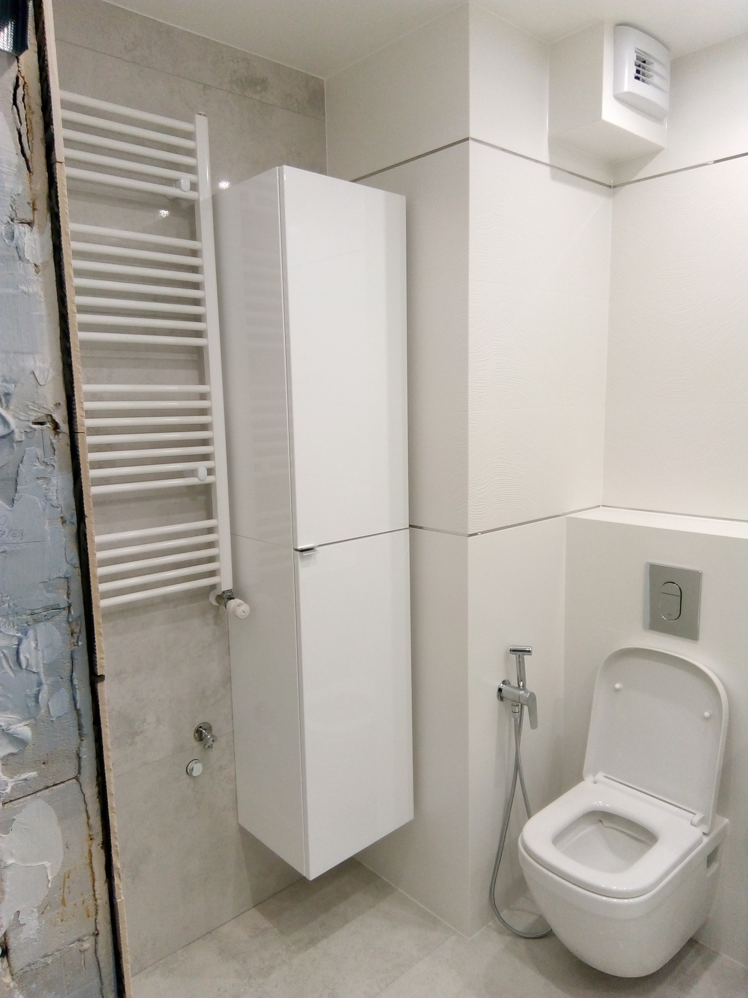 Nowoczesna, minimalistyczna łazienka z podwieszaną toaletą z bidetką, grzejnikiem drabinkowym i wysoką szafką wiszącą, widoczny fragment ściany przed wykończeniem.