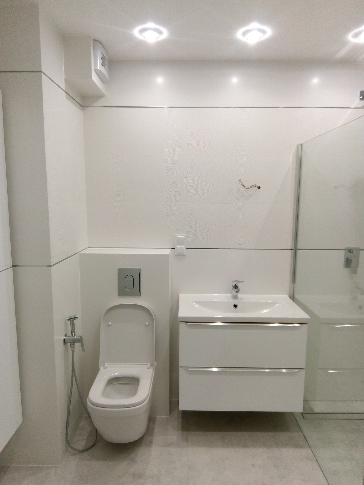 Nowoczesna, jasna łazienka z białą armaturą: wisząca toaleta z bidetką, umywalka z szafką i kabina prysznicowa ze szklanymi drzwiami, oświetlenie punktowe.