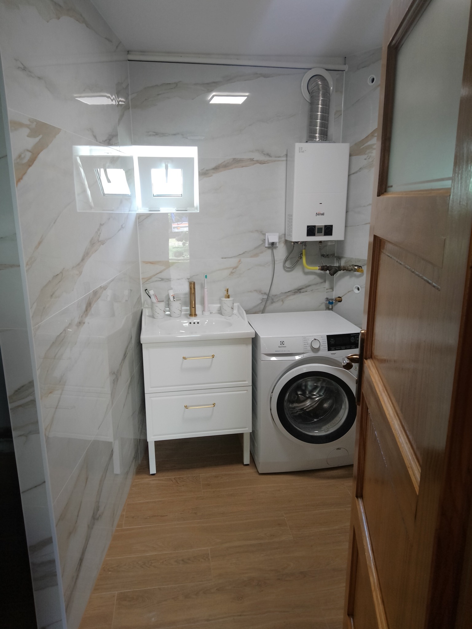 Nowoczesna łazienka z marmurowymi płytkami, umywalką z szafką, pralką Electrolux i piecem gazowym Ferroli, widok zza drewnianych drzwi.