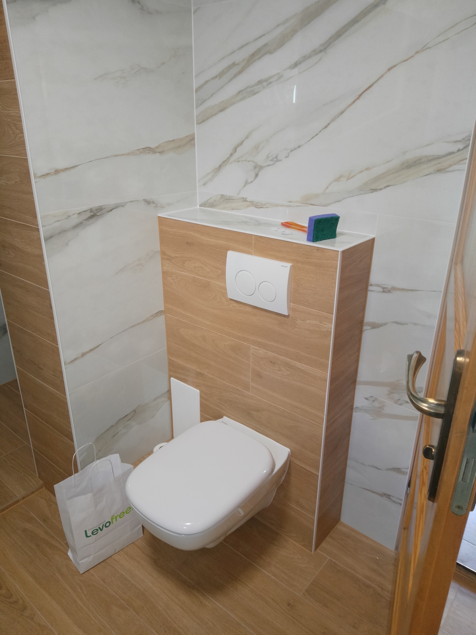 Zabudowana toaleta podtynkowa z białym przyciskiem spłukującym, wykończona płytkami imitującymi drewno, na tle marmurowych płytek ściennych. Na obudowie leży gąbka do czyszczenia, a obok toalety...