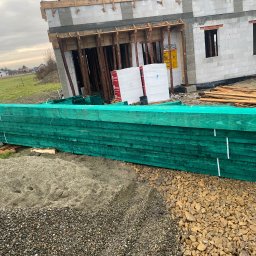 F.H.U OGIEL-DREW - Zielone, impregnowane deski na placu budowy, z surowym budynkiem w tle. Widoczne materiały budowlane i elementy konstrukcyjne.