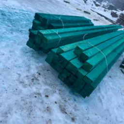 Impregnowane na zielono kantówki ułożone w stosy na śniegu. Widoczne ślady na śniegu i inne stosy drewna w tle. Drewno przygotowane do budowy.