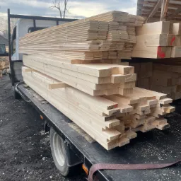 Świeżo przetarte deski i kantówki z jasnego drewna ułożone na pace samochodu dostawczego, widoczne czerwone oznaczenia na końcach niektórych elementów.