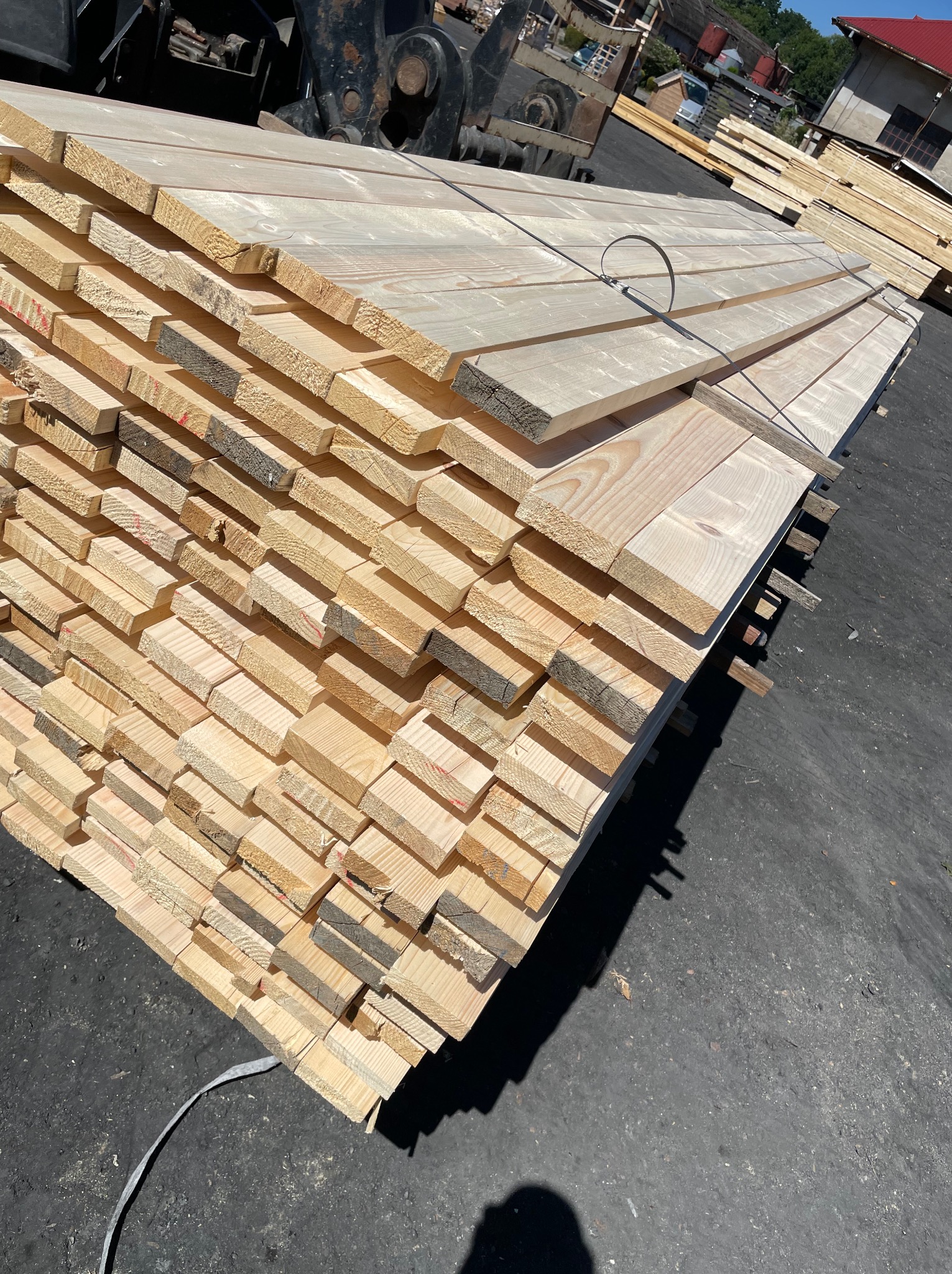 Stos tarcicy z jasnego drewna, zabezpieczony metalową taśmą, ułożony na zewnątrz. Widoczne słoje drewna i niewielkie przebarwienia. W tle budynki i fragment pojazdu.