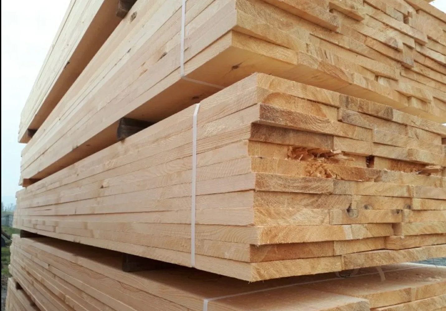 Ułożone w stosy deski z jasnego drewna, zabezpieczone białymi taśmami. Widoczne słoje drewna i niewielkie uszkodzenia na niektórych deskach. Tło rozmyte.