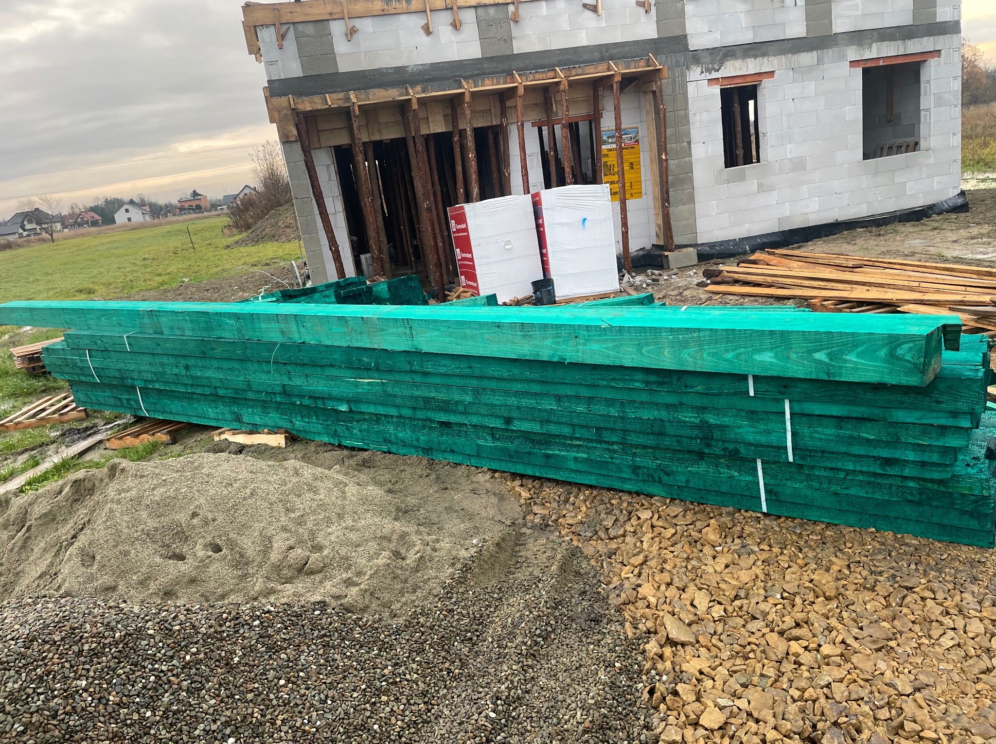 Zielone, impregnowane deski na placu budowy, z surowym budynkiem w tle. Widoczne materiały budowlane i elementy konstrukcyjne.