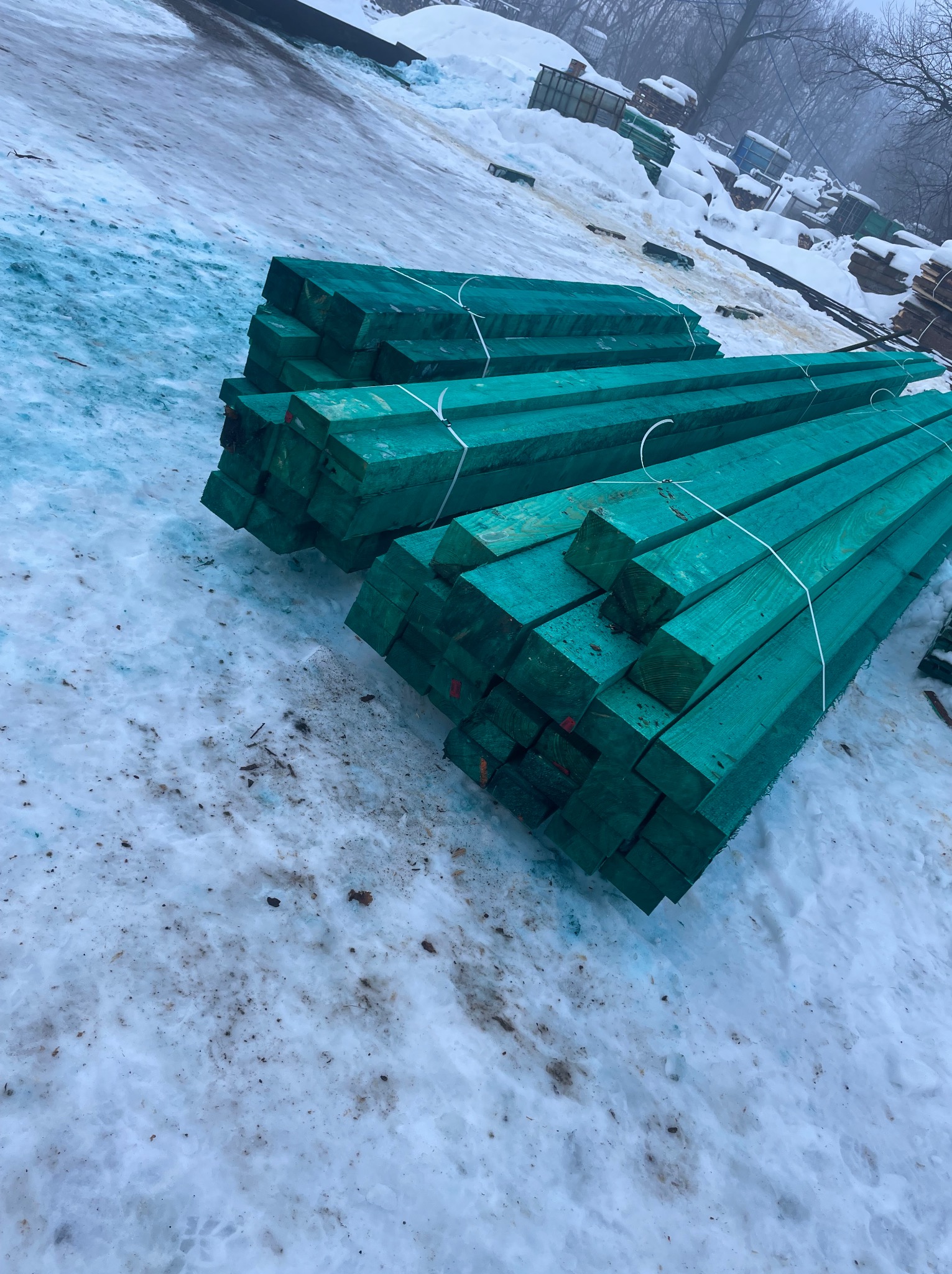 Impregnowane na zielono kantówki ułożone w stosy na śniegu. Widoczne ślady na śniegu i inne stosy drewna w tle. Drewno przygotowane do budowy.