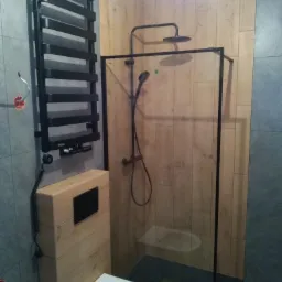 Nowoczesna łazienka z kabiną prysznicową typu walk-in z czarną ramą, grzejnikiem drabinkowym w kolorze czarnym i toaletą wiszącą z zabudową w drewnopodobnym dekorze.