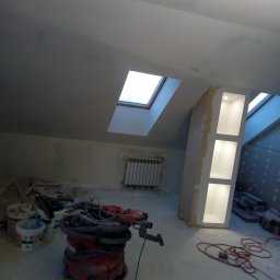 Stel-Bud - Poddasze w trakcie prac wykończeniowych: szpachlowane ściany, zabudowa z oświetlonymi niszami, okno dachowe, grzejnik, oraz narzędzia budowlane, w tym odkurzacz przemysłowy i pojemniki z zaprawą.