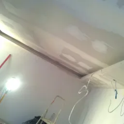 Sufit w trakcie szpachlowania z widocznymi łatami, drabina, lampa halogenowa i przewody elektryczne zwisające z sufitu.