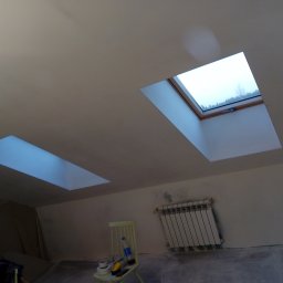 Stel-Bud - Poddasze po remoncie w Buczku z dwoma oknami dachowymi, widoczny grzejnik i krzesło z narzędziami malarskimi, ślady szpachlowania na ścianach.