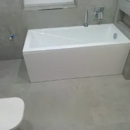 Nowoczesna łazienka z białą prostokątną wanną, szarą glazurą ścienną i podłogową oraz widoczną instalacją wodną na ścianie.