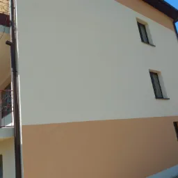 Ocieplona elewacja domu dwukondygnacyjnego z balkonem, pomalowana na dwa odcienie beżu, z widocznymi oknami i rynną, w słoneczny dzień.