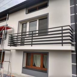 Stel-Bud - Elewacja domu po malowaniu z szarymi akcentami, widoczne rusztowanie i pracownik przy pracy, balkon z czarną balustradą, okna w drewnianych ramach.