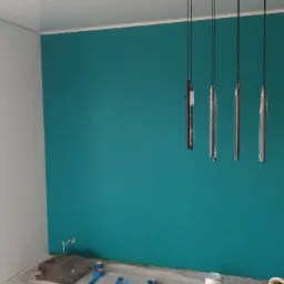 Świeżo pomalowane wnętrze: turkusowa ściana z wiszącymi lampami w ochronnej folii, obok fragment białej ściany, widoczne akcesoria malarskie przy podłodze.