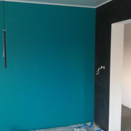 Stel-Bud - Ściana pomalowana na turkusowo z wiszącymi lampami w folii ochronnej, kontrastująca z czarną ścianą i białym obramowaniem drzwi prowadzących do pomieszczenia w trakcie prac wykończeniowych.