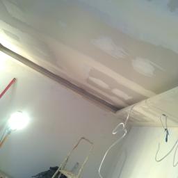 Stel-Bud - Sufit w trakcie prac wykończeniowych, widoczne płyty karton-gips z nałożoną warstwą szpachli, drabina i lampa robocza w rogu pomieszczenia, przewody elektryczne zwisające z sufitu.