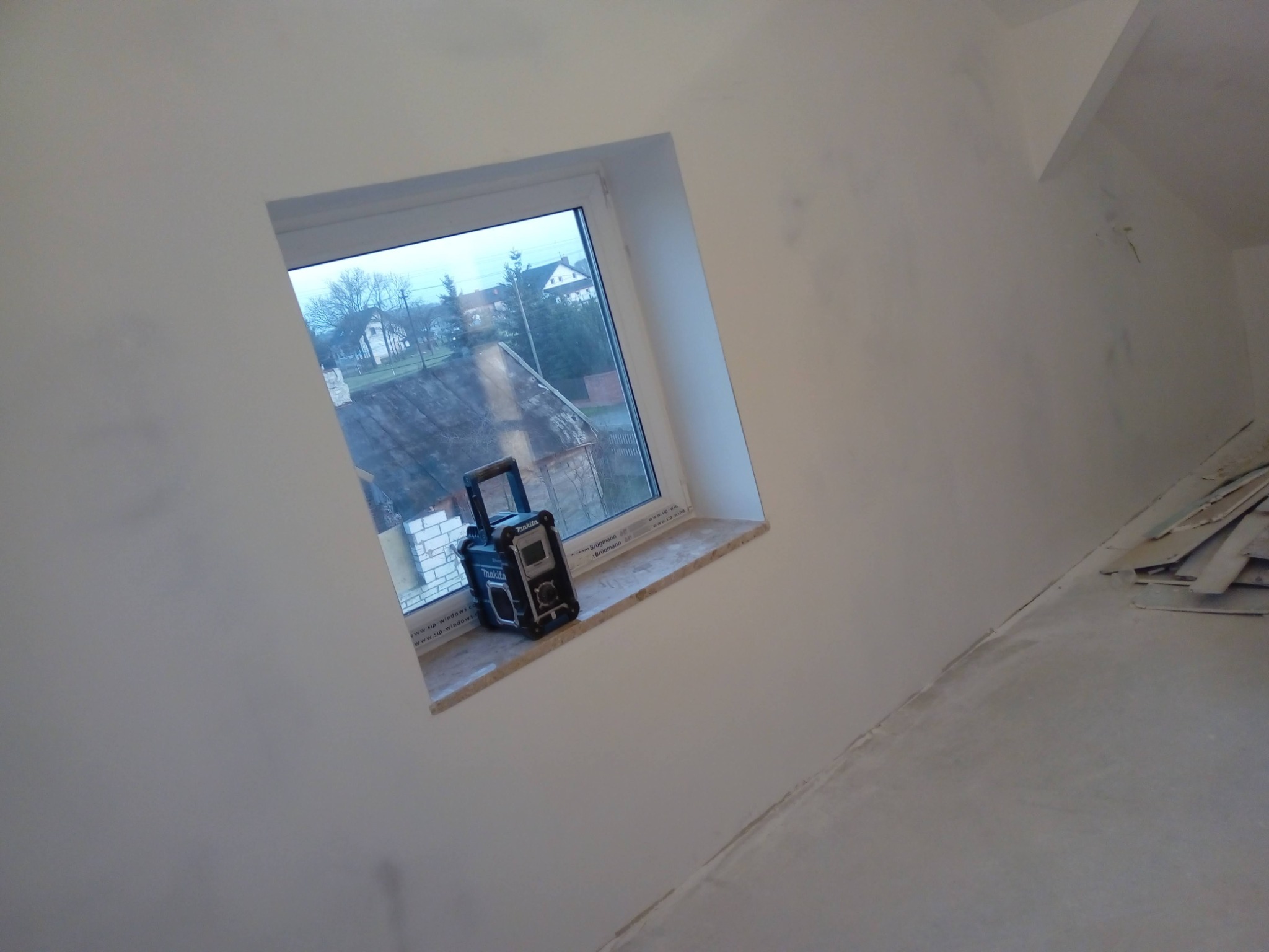 Poddasze w trakcie prac wykończeniowych: ściany po nałożeniu gładzi, widoczne niedoskonałości i nierówności, radio Makita na parapecie okna z widokiem na zewnątrz.