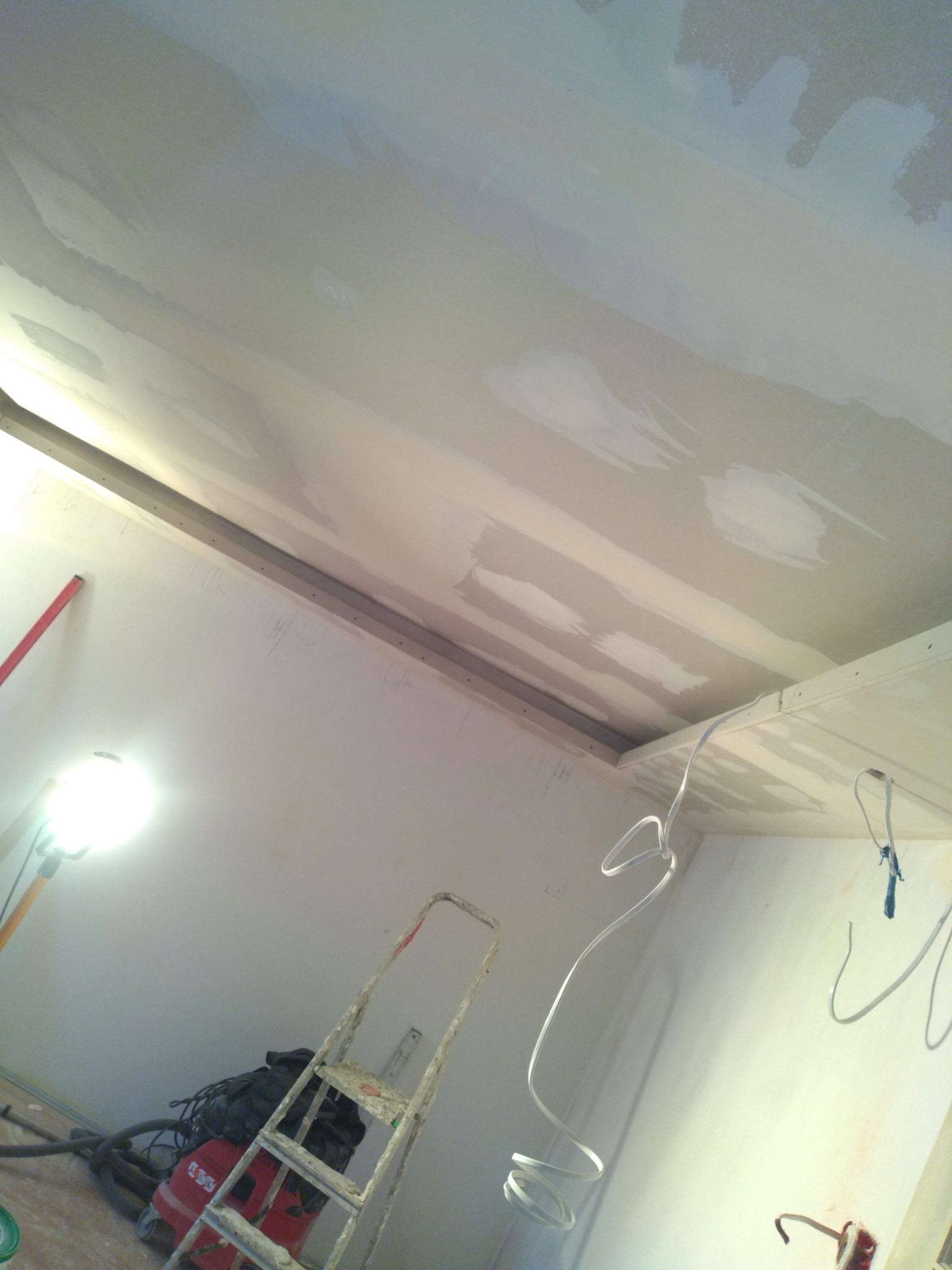 Sufit w trakcie szpachlowania z widocznymi łatami, drabina, lampa halogenowa i przewody elektryczne zwisające z sufitu.