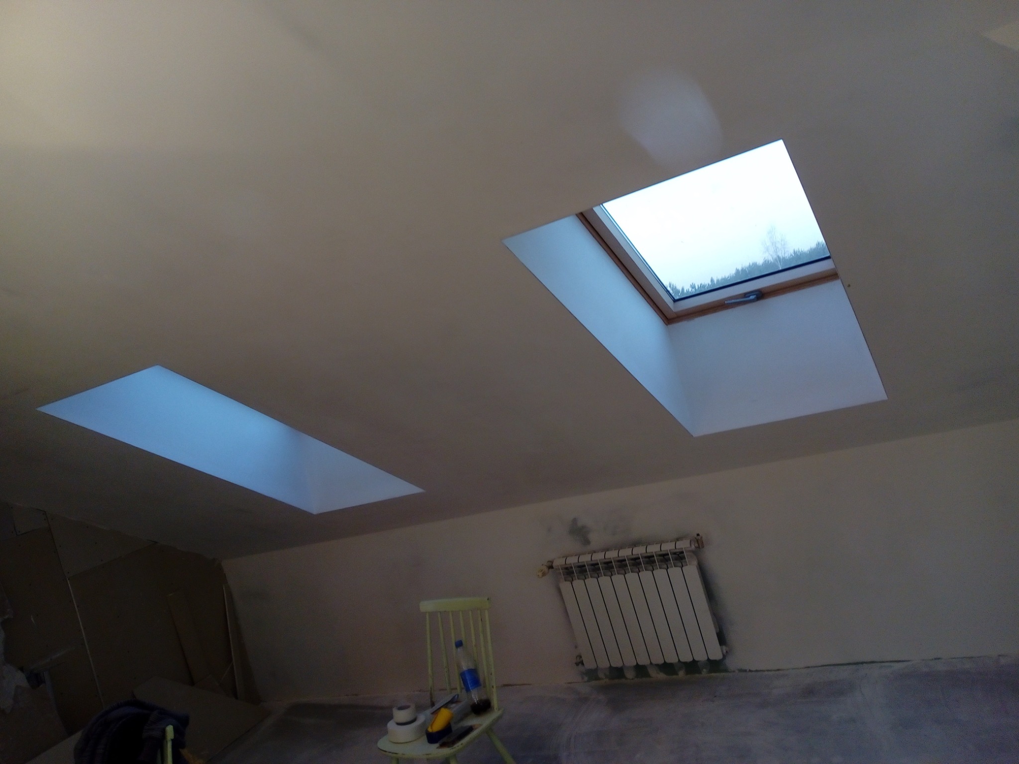 Poddasze po remoncie w Buczku z dwoma oknami dachowymi, widoczny grzejnik i krzesło z narzędziami malarskimi, ślady szpachlowania na ścianach.