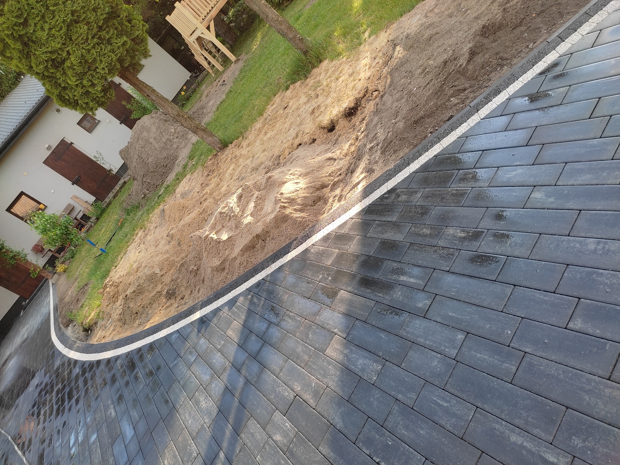 Ułożona, ciemnoszara kostka brukowa w Borowie, wykończona jasną obwódką, przylegająca do ziemi przygotowanej pod trawnik. Widoczny fragment budynku w tle.