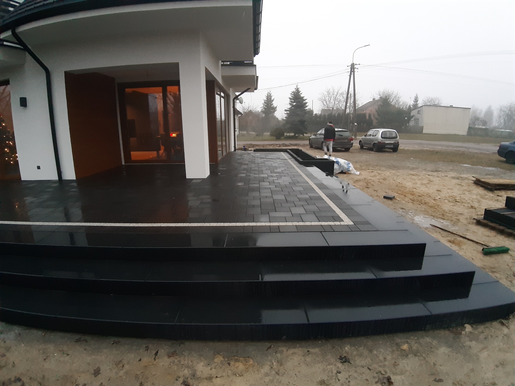 Nowoczesny taras z ciemnej kostki brukowej i schodami, z jasną obwódką, widok na dom z dużymi oknami i choinką w środku, częściowo ukończone prace, w tle zaparkowane samochody i osoba.